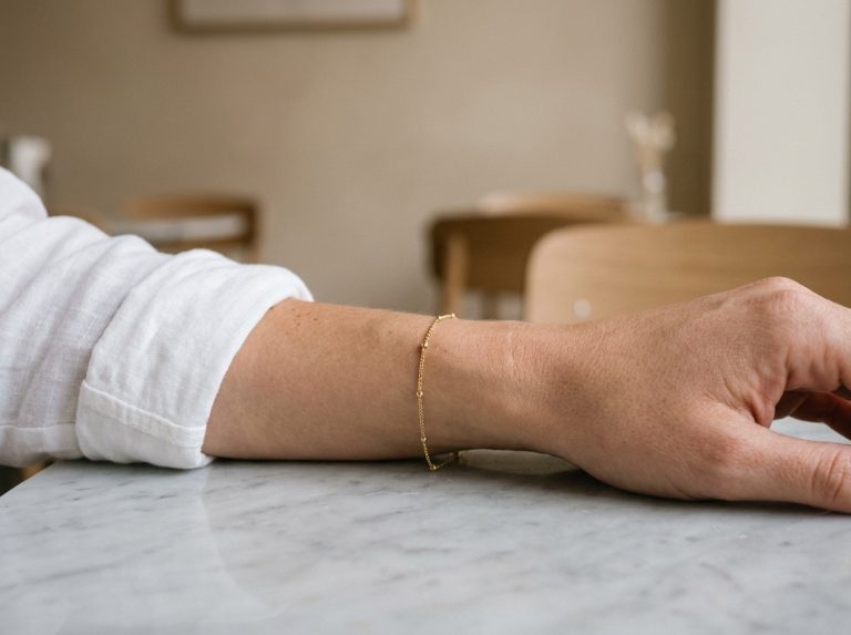 Bracelet fin doré porté au poignet d'une jeune femme en chemise blanche, posé sur une surface en marbre dans un café minimaliste
