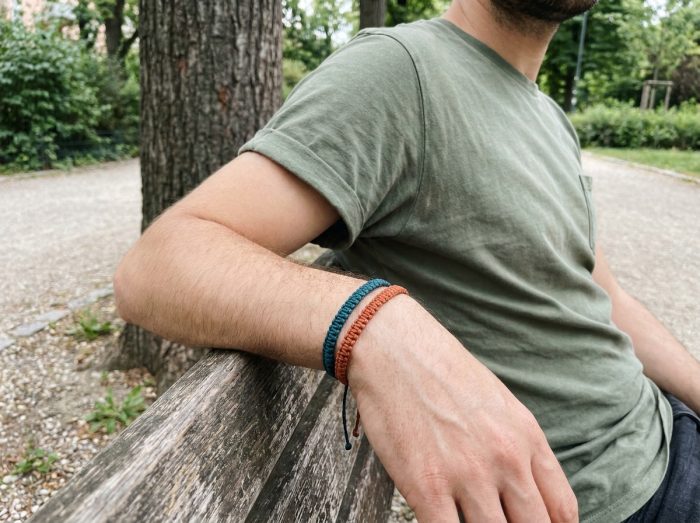 Bracelets en cordon coloré teal et terracotta portés par un homme en t-shirt kaki assis sur un banc en bois dans un parc urbain