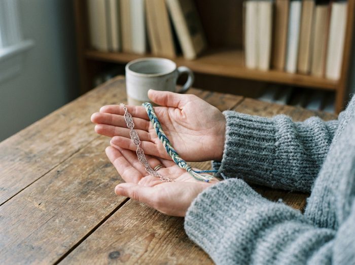 Femme tenant un bracelet fin argenté et un cordon tressé multicolore dans ses mains pour les comparer, ambiance intérieur cosy avec pull gris